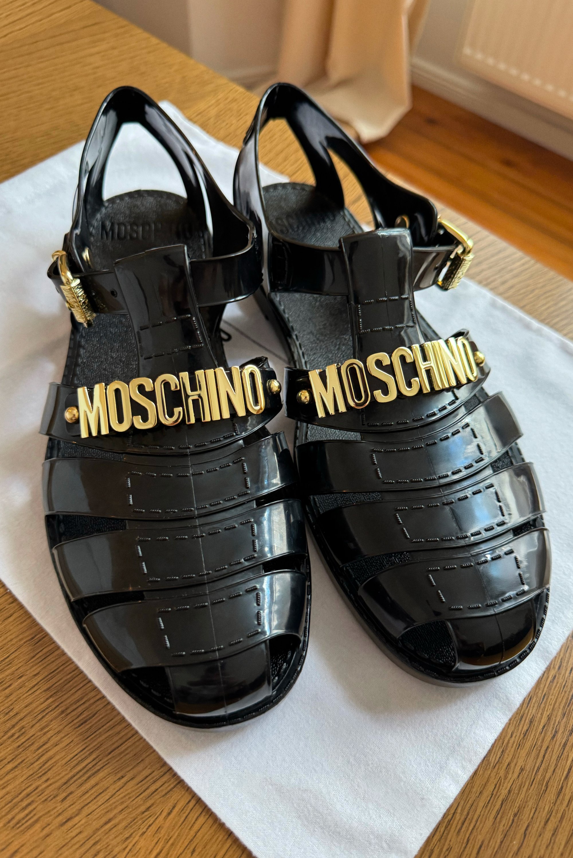 Sandali in gel nero con superficie texturizzata, caratterizzati dal branding dorato "Moschino", design open-toe e cinturino alla caviglia con fibbia dorata.