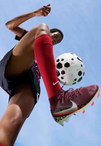 Joueur de football portant des chaussettes et des crampons Nike rouges frappe un ballon de football noir et blanc sur fond de ciel bleu clair.