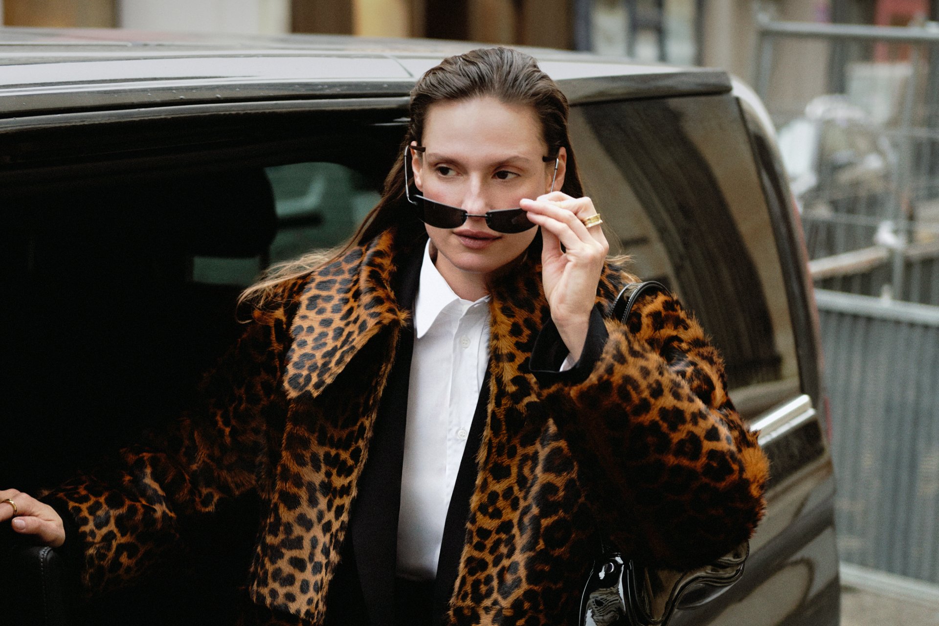 A woman in a leopard-print coat adjusts her sunglasses while exiting a black vehicle, showcasing a confident urban fashion moment.
