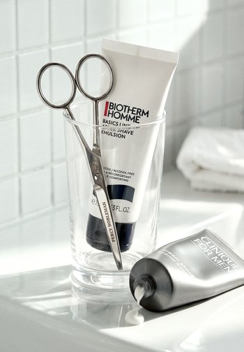 Tubetti di emulsione da barba e forbici in un bicchiere trasparente su un piano da bagno piastrellato bianco con una piccola quantità di crema versata.