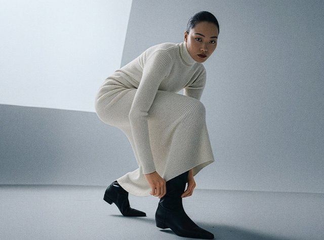 Ribbstickad krämfärgad klänning med hög hals och långa ärmar, kombinerad med svarta ankelboots, mot en minimalistisk bakgrund.
