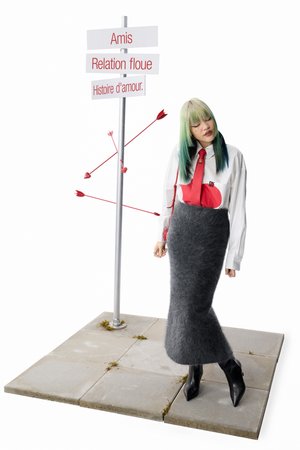 Una persona con el pelo de puntas verdes se encuentra junto a un cartel etiquetado con amistad, relaciones inciertas y amor, rodeada de flechas con corazones.