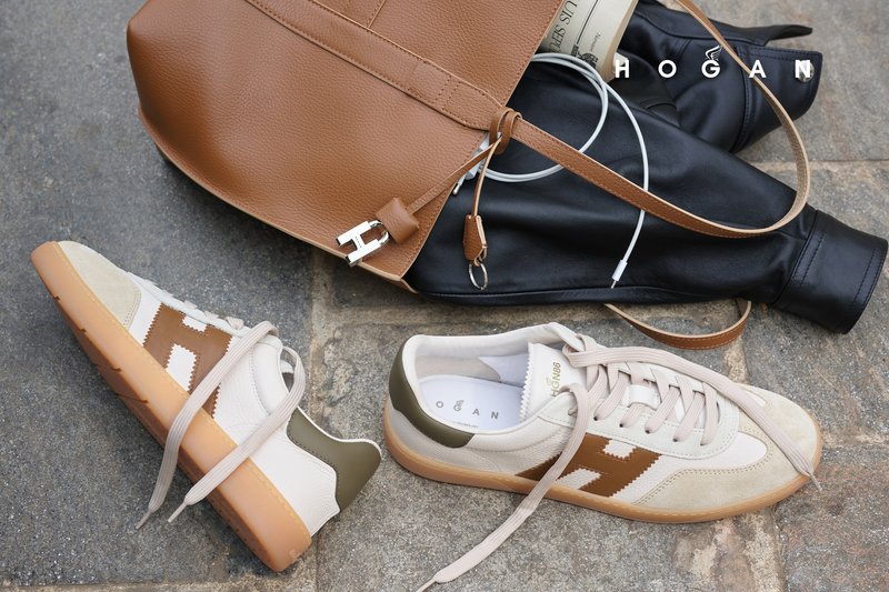 Paar beige-braune Hogan-Sneaker neben einer hellbraunen Lederhandtasche und einer schwarzen Lederjacke auf einem Steinboden mit weißen Ohrhörern.