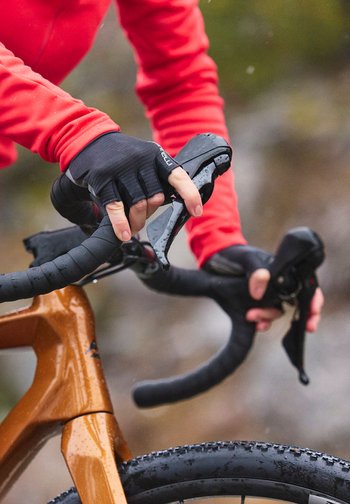 Hender i svarte hansker som holder sykkelstyret, iført en rød langermet jakke, med en brun sykkelramme og vått terreng i bakgrunnen.