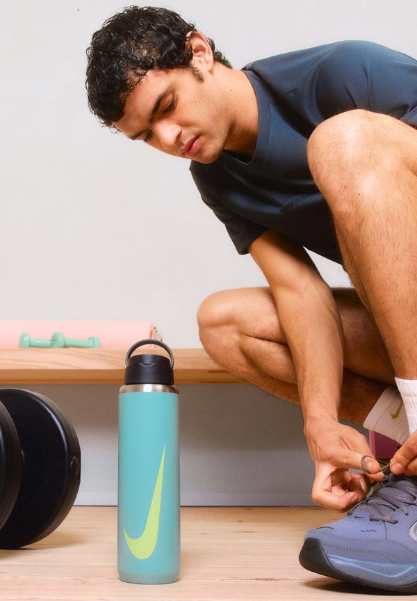 Nerezová oceľová fľaša na vodu v tyrkysovej farbe so zeleným logom Nike. Atlet má na sebe tmavé tričko a sivé topánky, viaže si šnúrky na drevenom povrchu.