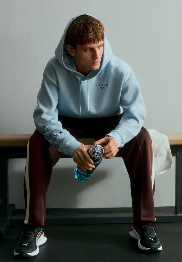 Young man in light blue hoodie and maroon pants sitting on bench, holding a water bottle, looking to the side in a minimal indoor setting.
