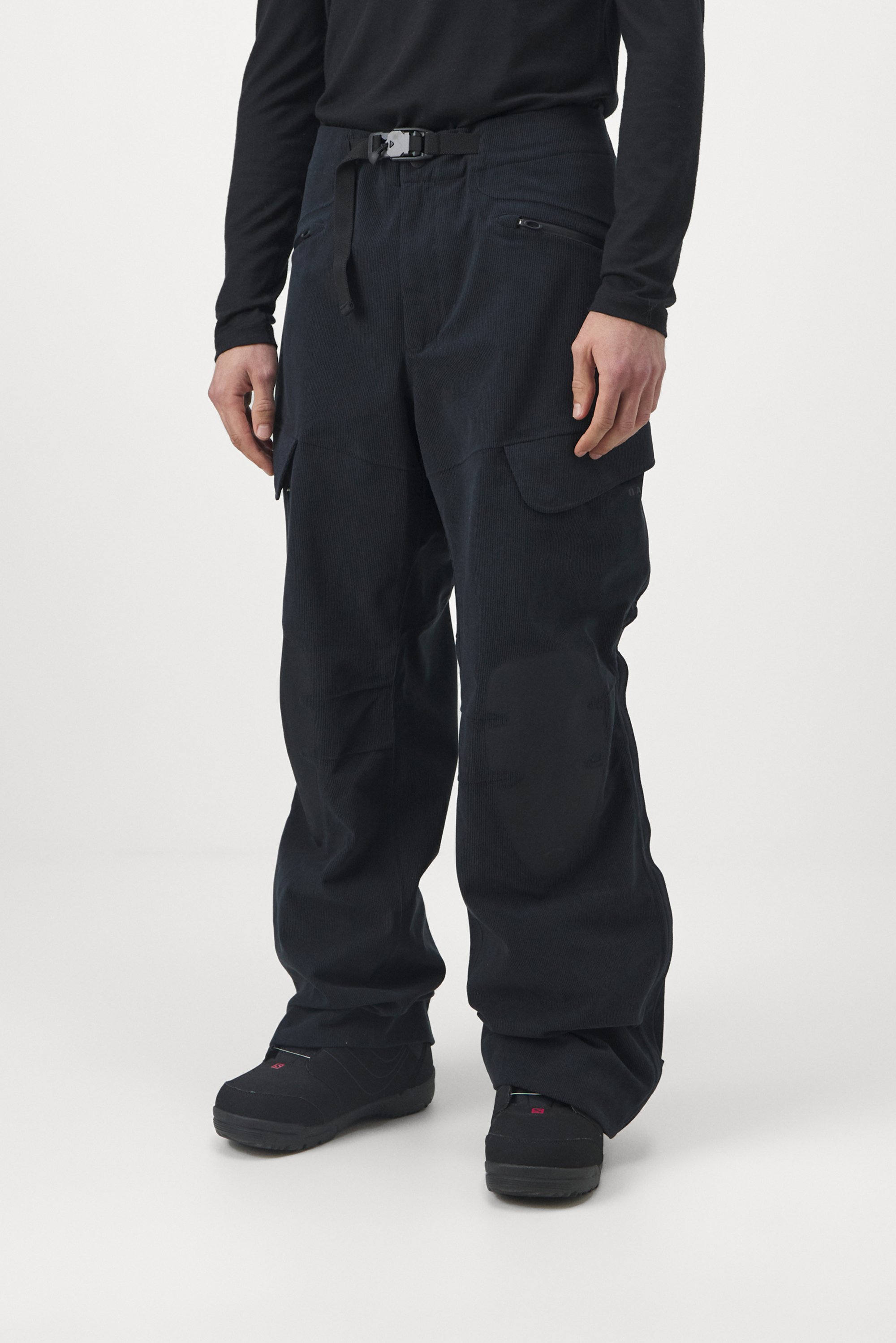 Man wearing dark cargo pants with multiple pockets and a belt, paired with dark shoes, against a minimalistic white background.