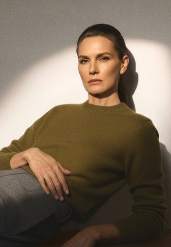 Olivengrønn, strikket genser med rund hals. Modellen har hånden sin på grå bukser, minimalistisk bakgrunn med mykt lys og skygger.