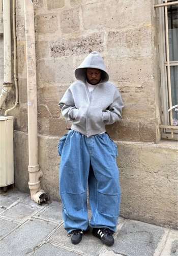 Uma hoodie cinza com fecho de correr e um capuz grande, combinada com calças de ganga azul folgadas com bolsos laterais. Ténis pretos com riscas brancas.
