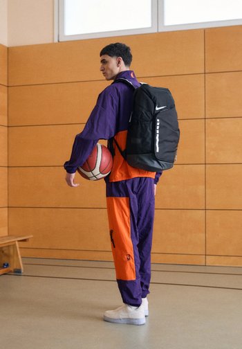 Un jeune homme en tenue de sport violette et orange tient un ballon de basket et porte un sac à dos noir Nike dans une salle de sport.