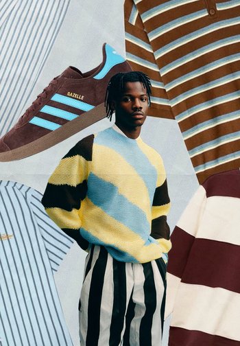 Barna velúr sneakers kék díszítéssel. A modellen csíkos sárga, kék és barna kötött pulóver van, amit fekete-fehér csíkos nadrággal párosítottak.