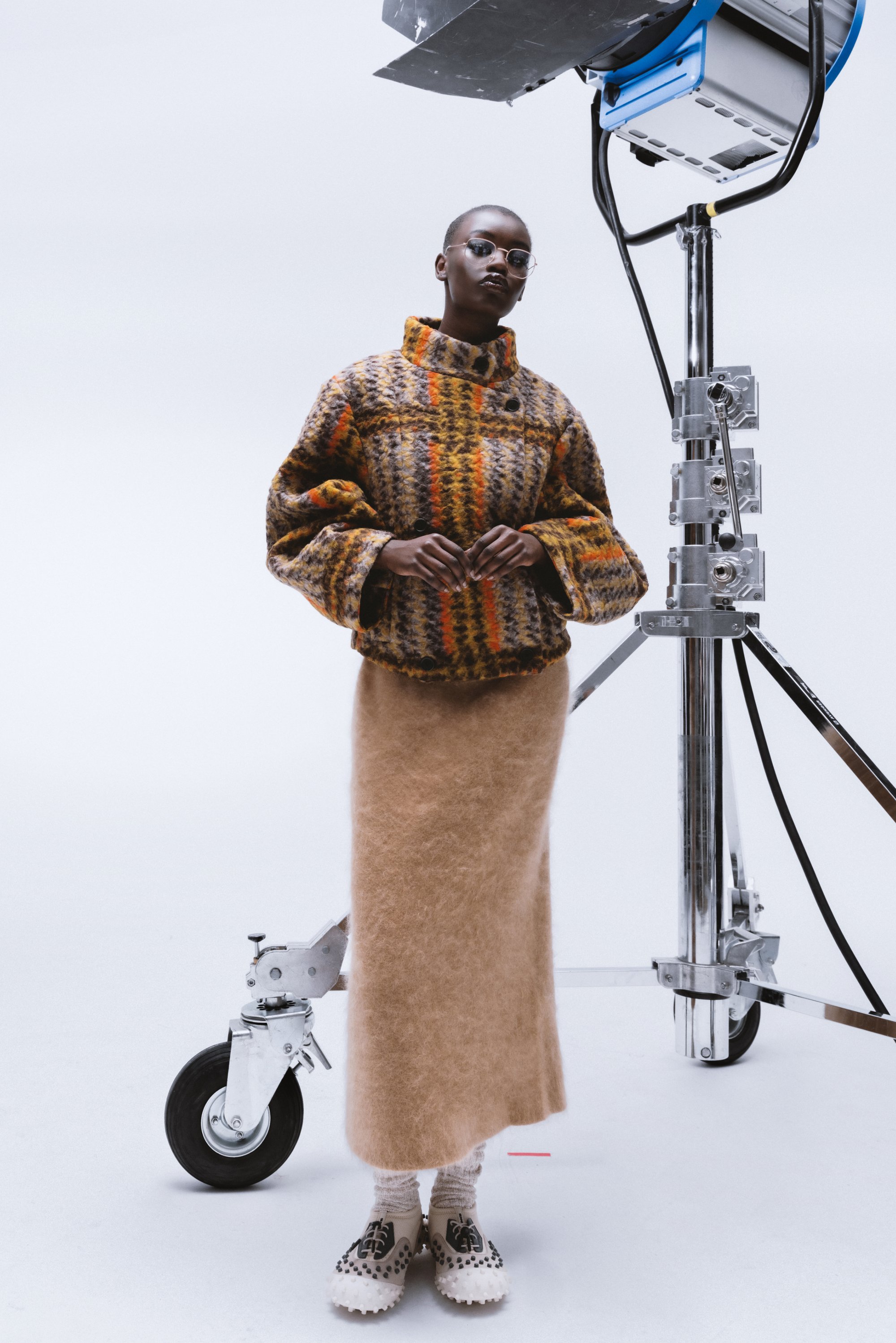Modelo con un suéter oversized de estampado y una larga falda de terciopelo se encuentra cerca de una luz de cine. Lleva gafas y zapatos únicos, posando con confianza.