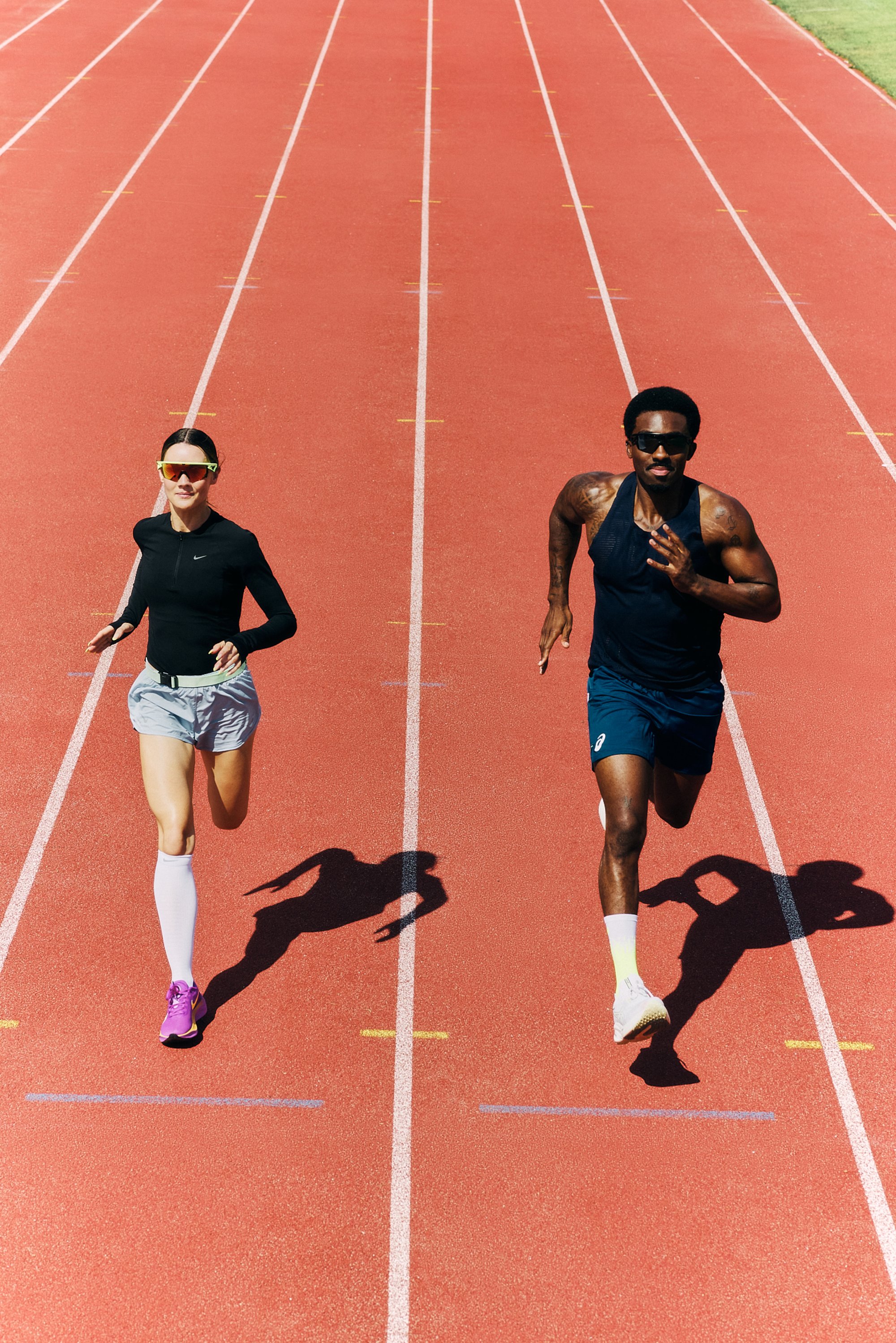 Two sprinting runners on a red track: a man in a sleeveless top and a woman in a long-sleeve shirt and shorts, both wearing sunglasses.