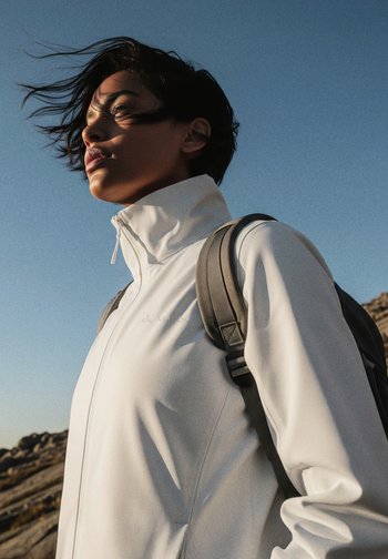 Giacca bianca con zip realizzata in materiale morbido e leggero, dotata di colletto alto e dettagli con logo discreto. Cinghia dello zaino grigia visibile.