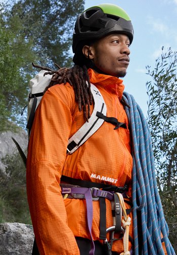 Man wearing green helmet and orange jacket, carrying blue climbing rope and backpack, standing outdoors near trees and rocks.