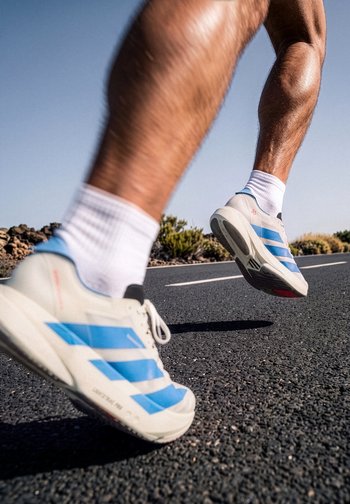 Runner wearing white shoes with blue stripes and white socks running on a paved road under clear sky.