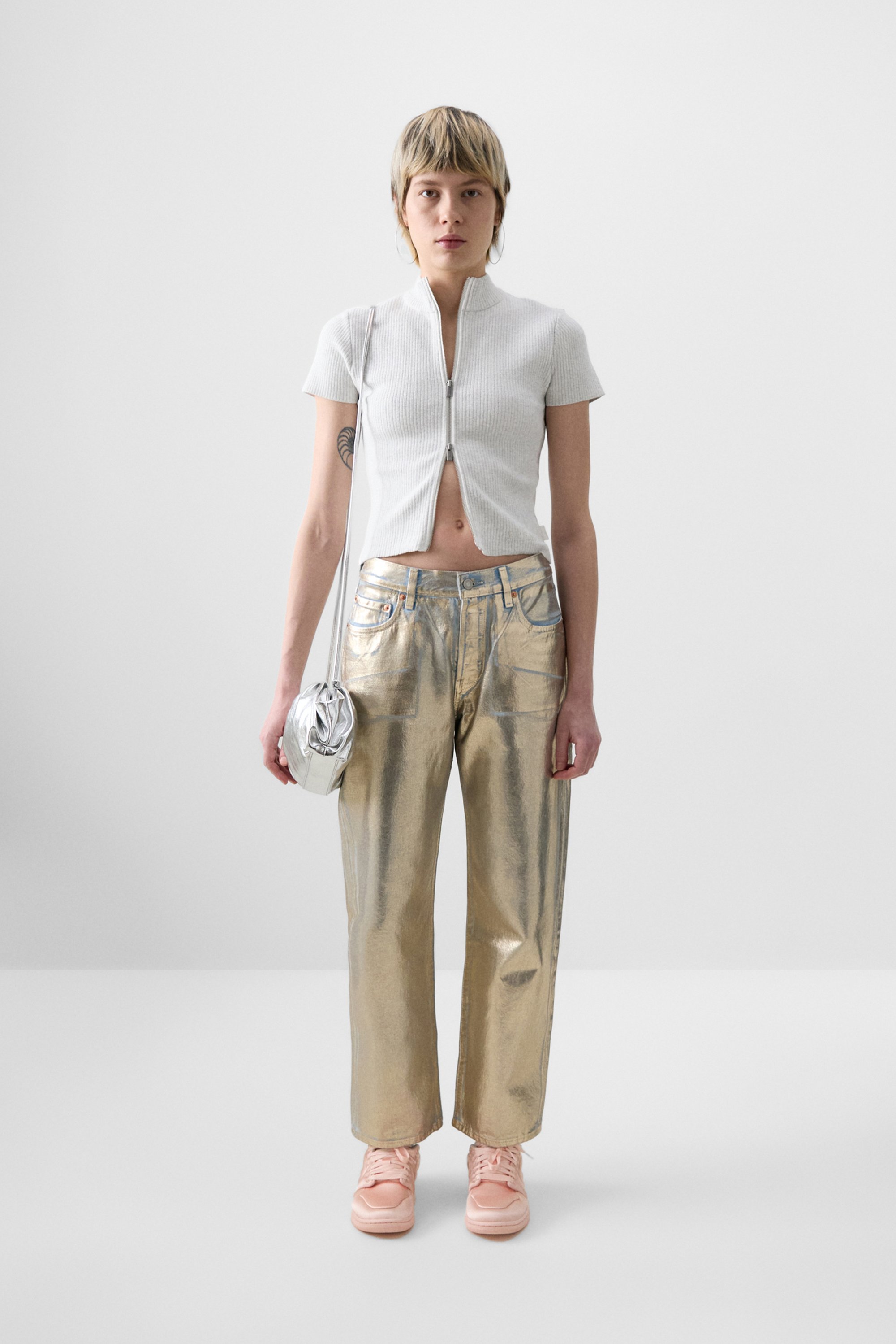 Una persona si trova davanti a uno sfondo neutro, indossando un top bianco con zip corto, pantaloni dorati lucidi e sneaker rosa, mentre tiene una piccola borsa argentata.