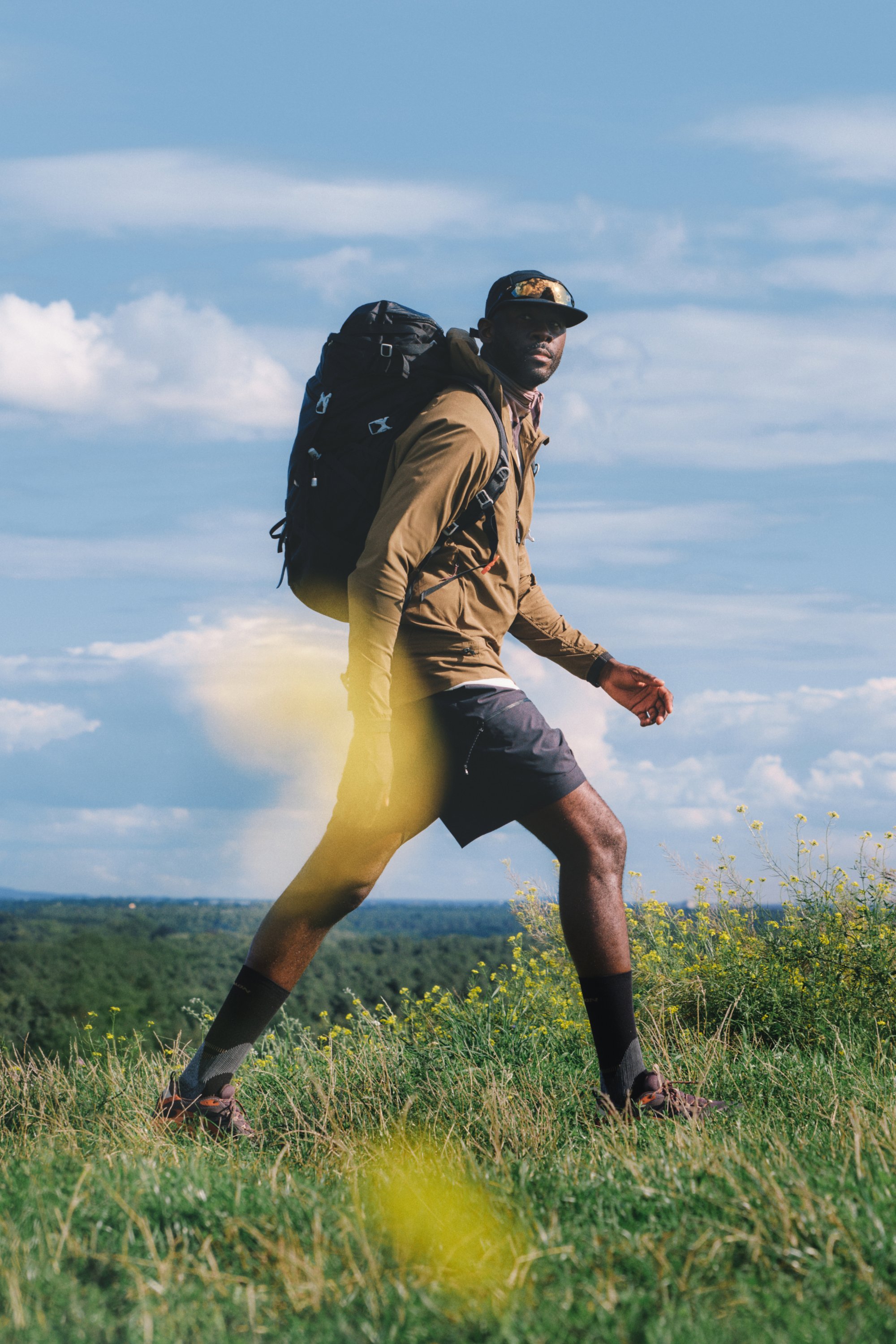 Zwarte rugzak met meerdere vakken, gedragen door een persoon in een beige jas en shorts, die door het groene gras loopt, omringd door gele bloemen.