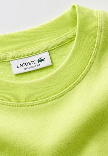 Sudadera verde neón con cuello redondo acanalado y etiqueta Lacoste que muestra un ajuste holgado y logotipo de cocodrilo verde.