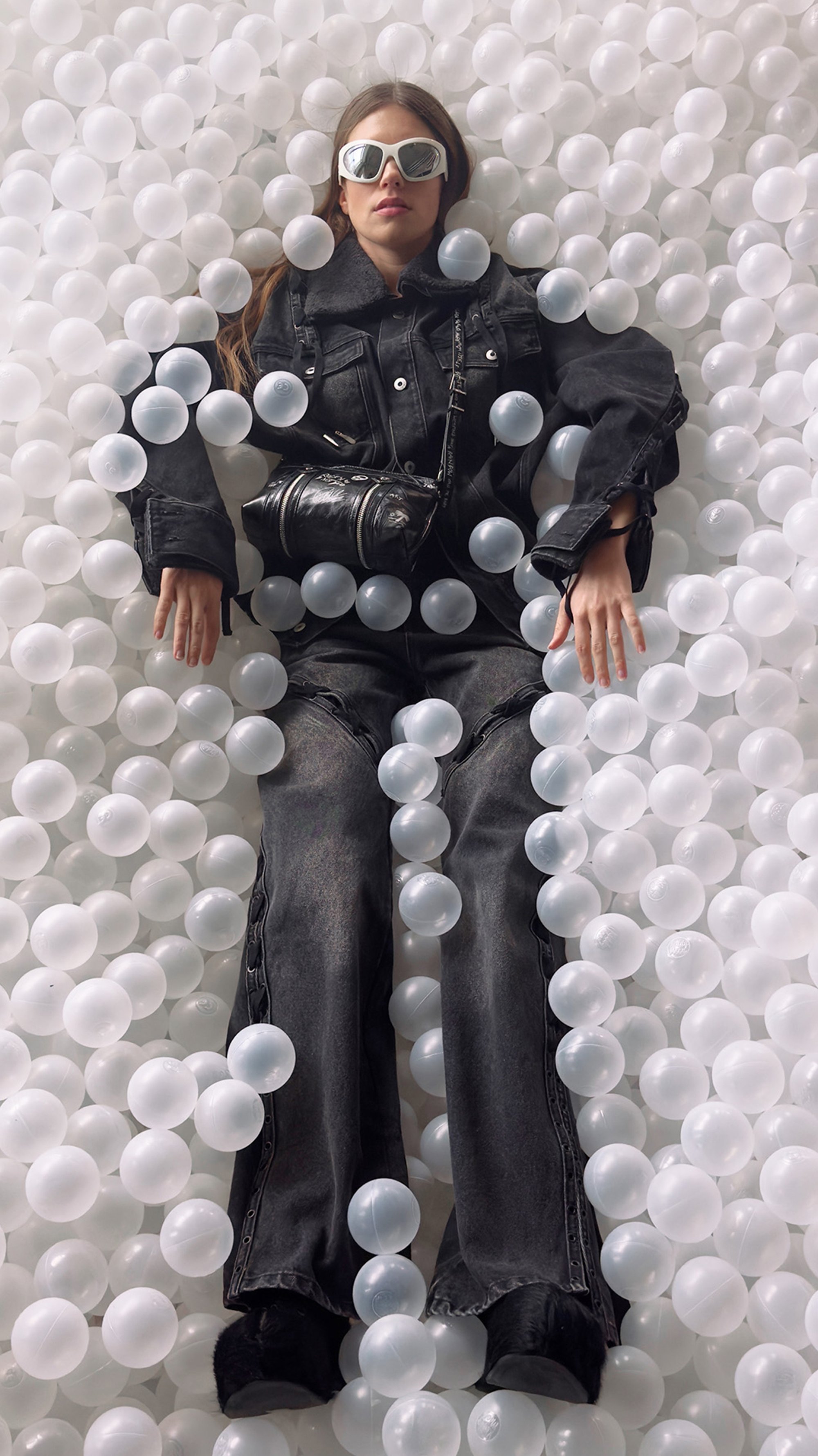 A person in sunglasses and a black outfit lies among scattered translucent balls, exuding a relaxed, playful vibe in a vibrant setting.