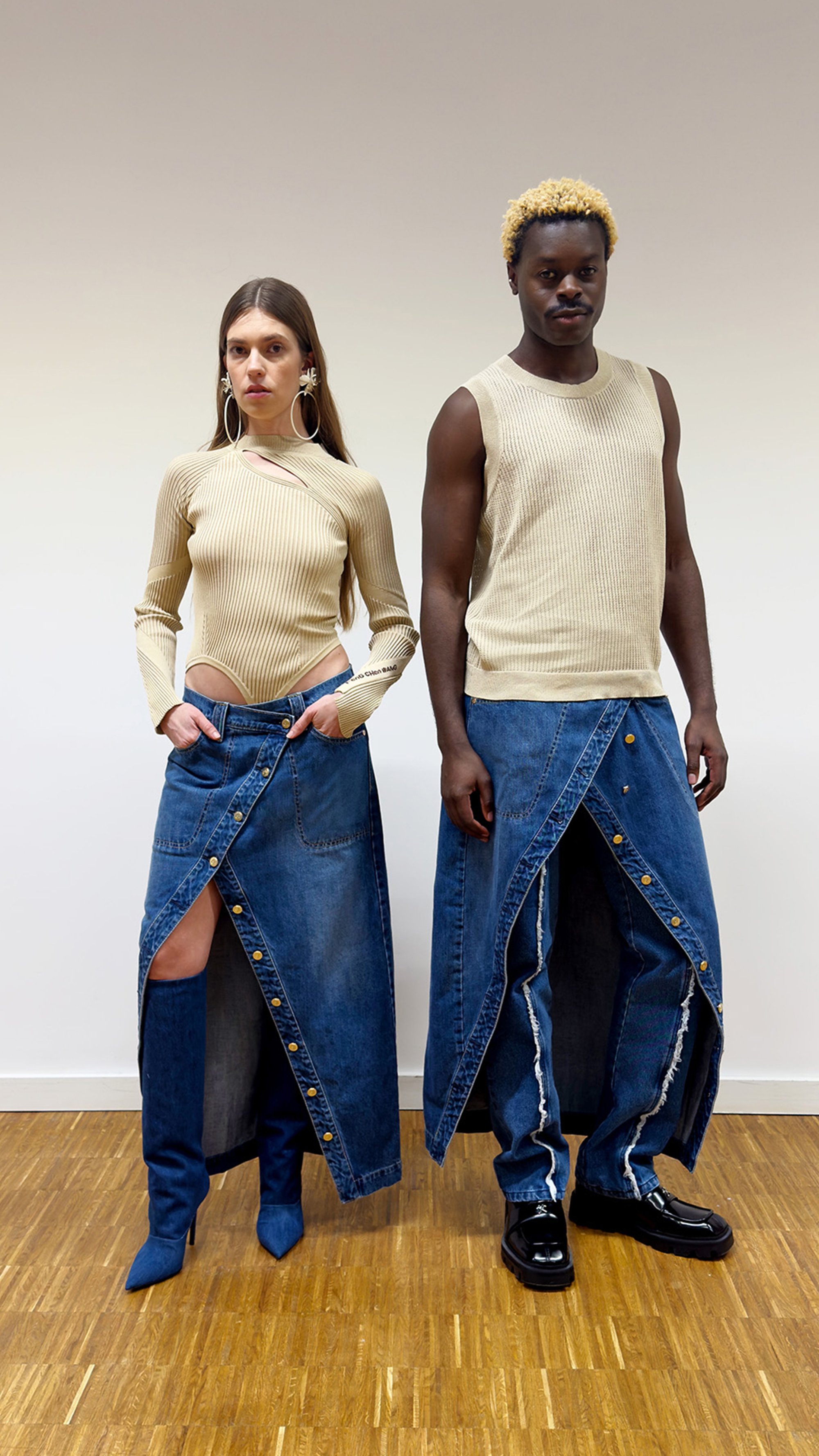 A woman in a beige top and a man in a sleeveless beige shirt stand side by side, both wearing denim skirts with buttons and high boots.