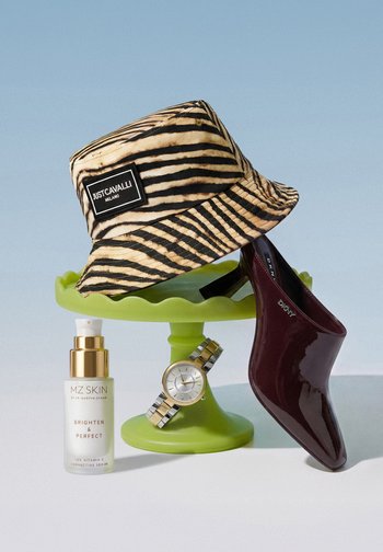 Striped Just Cavalli bucket hat, burgundy DKNY pointed shoe, silver and gold wristwatch, and MZ Skin serum on a green pedestal against pale blue background.