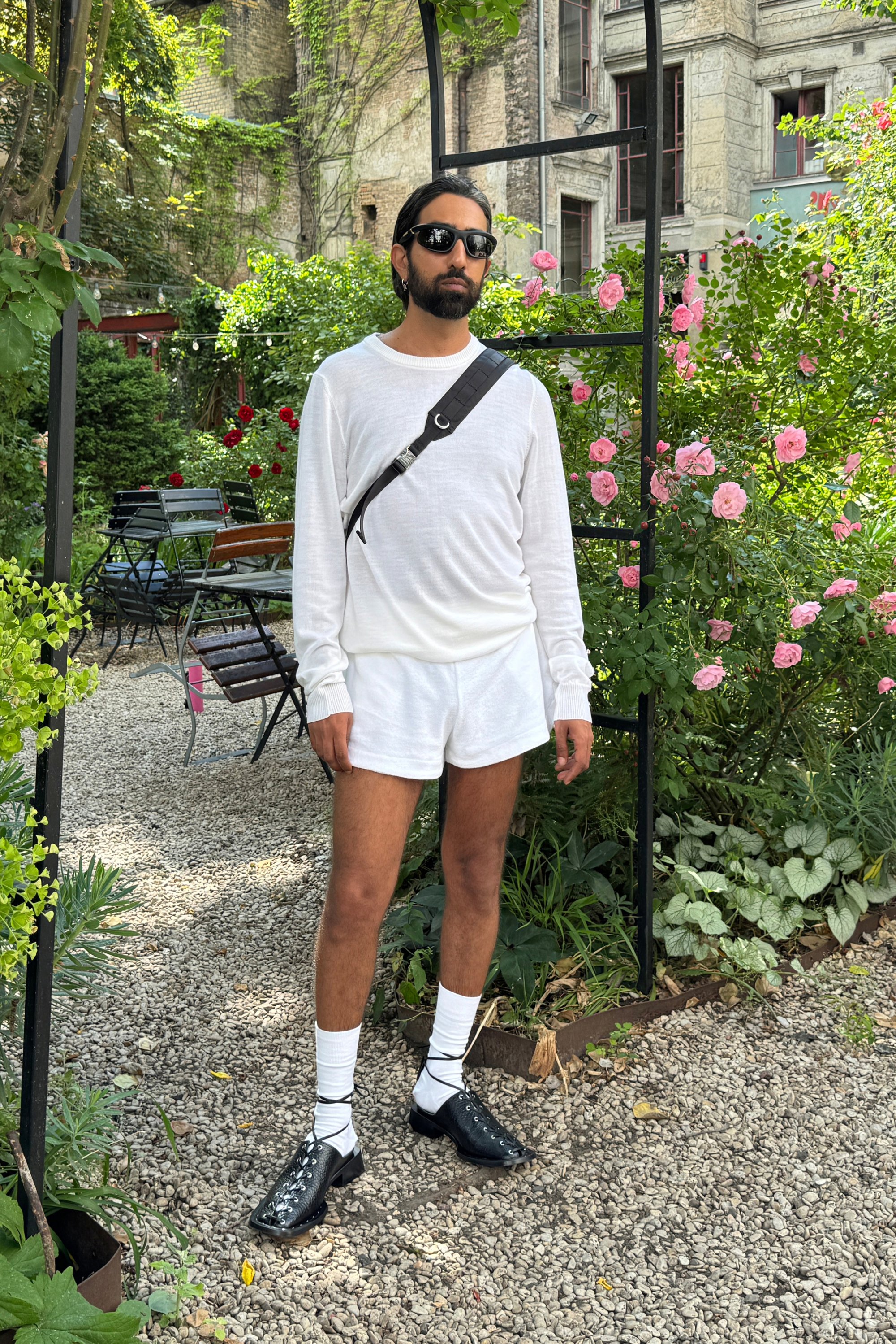 Un uomo è in piedi in un giardino, indossando una camicia bianca a maniche lunghe e pantaloni corti, scarpe nere e occhiali da sole, circondato da vegetazione e rose.