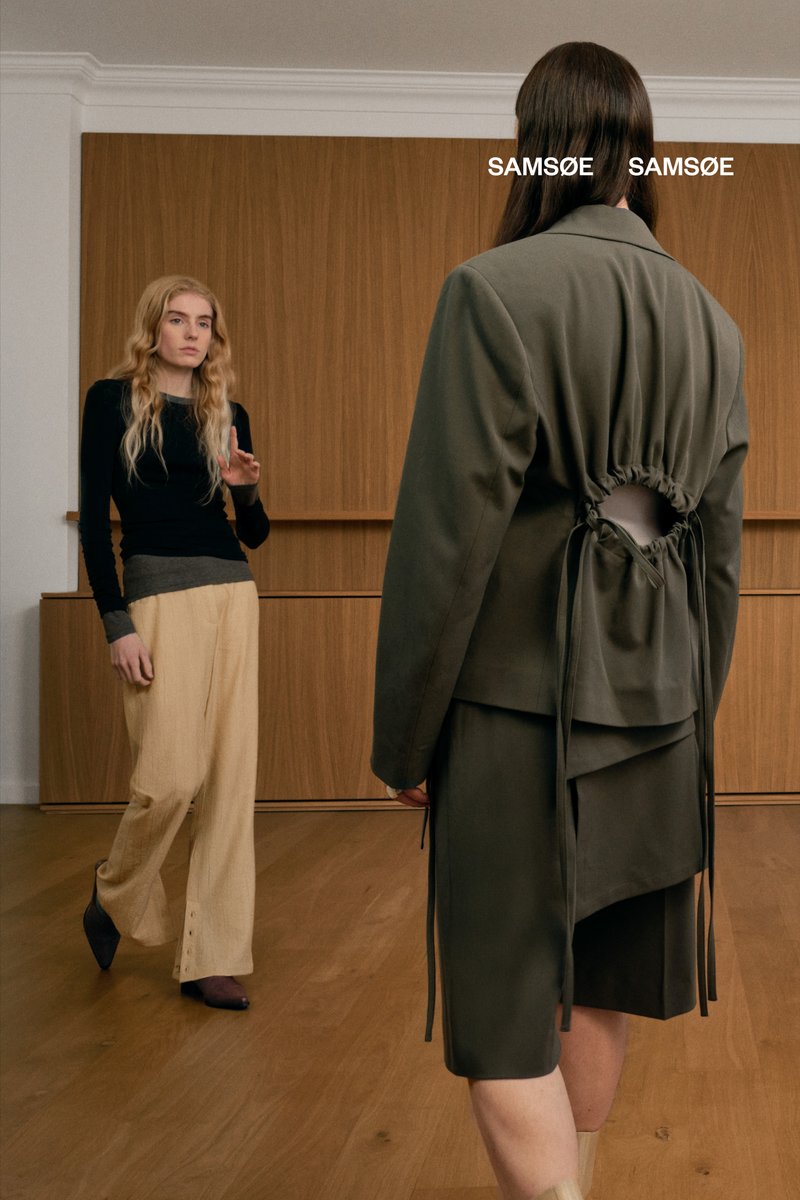 Dos personas en una habitación minimalista; una lleva una camiseta negra y pantalones beige, gesticulando, mientras que la otra viste un elegante conjunto verde con un corte en la espalda.