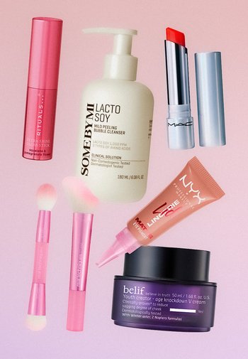 Roze en paarse cosmetische producten op een roze achtergrond, waaronder lipgloss, reiniger, lippenstift, oogpigment, crème en twee make-upkwasten.