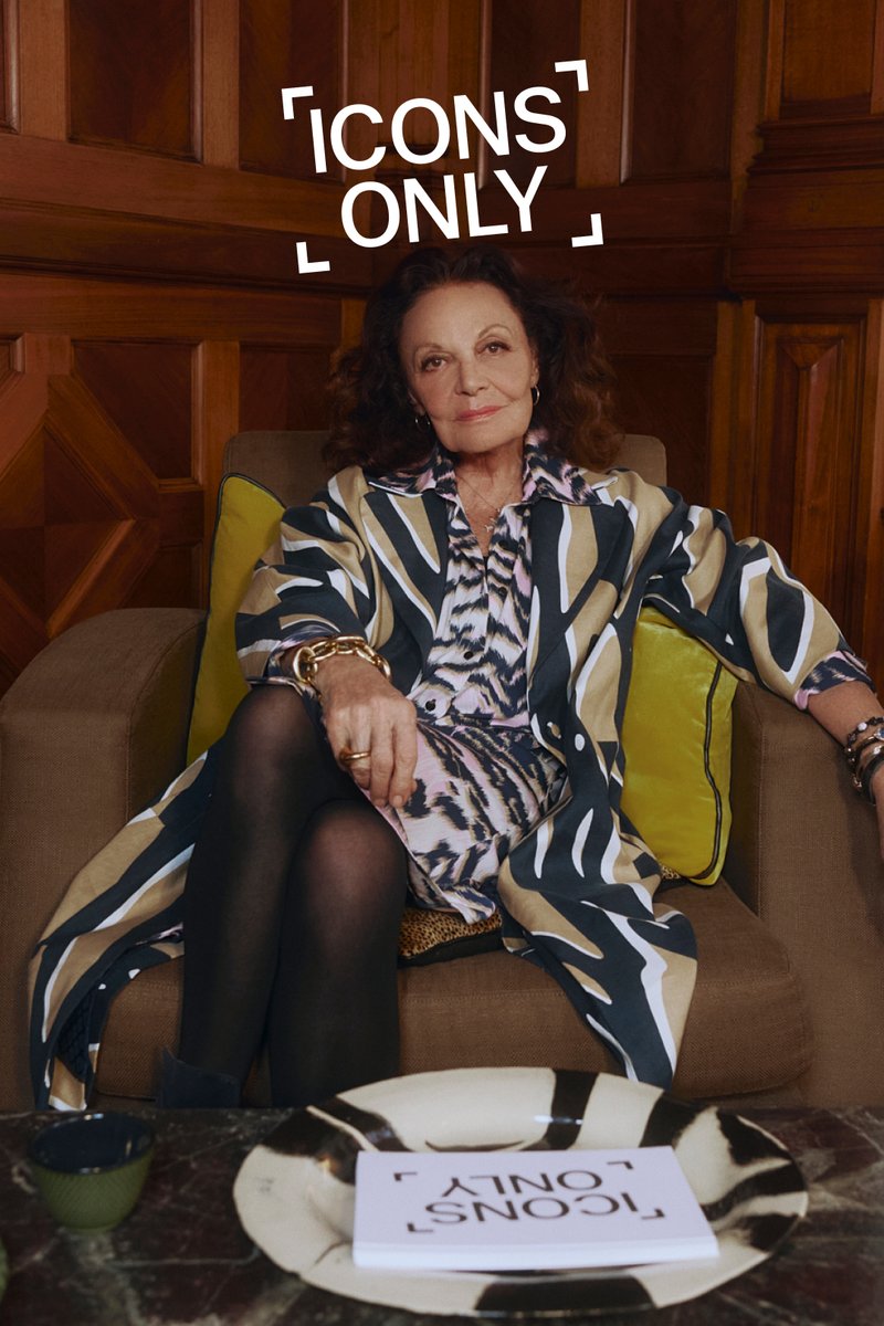 En kvinde med krøllet hår sidder på en sofa iført en mønstret kimono. En bog med titlen "IKONER KUN" ligger på et bord ved siden af hende.