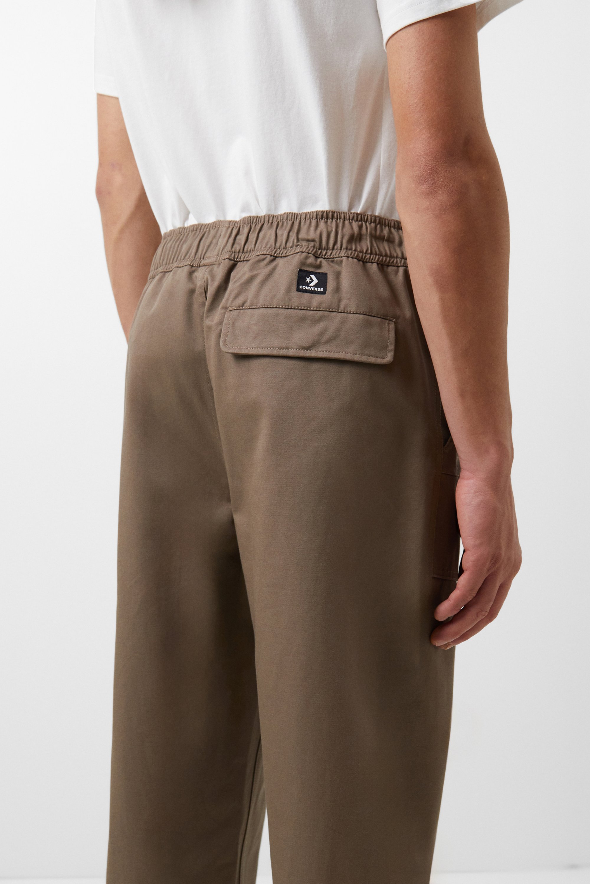 Een persoon in een wit shirt staat, toont de achterkant van een bruine broek met een elastische tailleband en een zak met een logo.