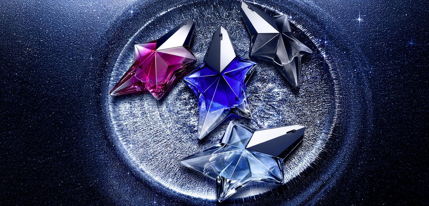 Fire stjerneformede parfumeflasker i pink, blå, klar og mørkegrå glas med facetterede overflader, udstillet mod en mørk, glitrende baggrund.