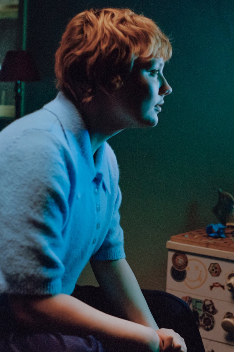 Personne aux cheveux courts portant une chemise bleue, assise et regardant intensément vers la droite dans une pièce faiblement éclairée près d'un tiroir avec des autocollants.