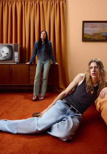 Junge Person mit langen Haaren lehnt sich auf einem orangefarbenen Teppich zurück; ältere Person mit langen dunklen Haaren lehnt an einem Mid-Century-Schrank, auf dem ein Außerirdischer im Fernseher zu sehen ist.