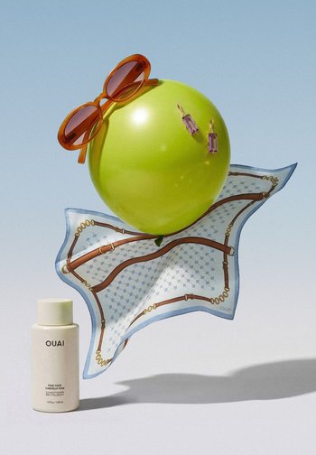Grüner Luftballon mit brauner Sonnenbrille und lila Ohrringen schwebt über einem gemusterten Seidenschal und einer Flasche OUAI Haarspülung.