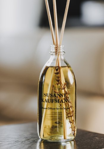 Szklana butelka z jasnożółtym płynem, trzema trzcinowymi patyczkami i suszonymi ziołami w środku, oznakowana "Susanne Kaufmann Room Diffuser for the Senses."