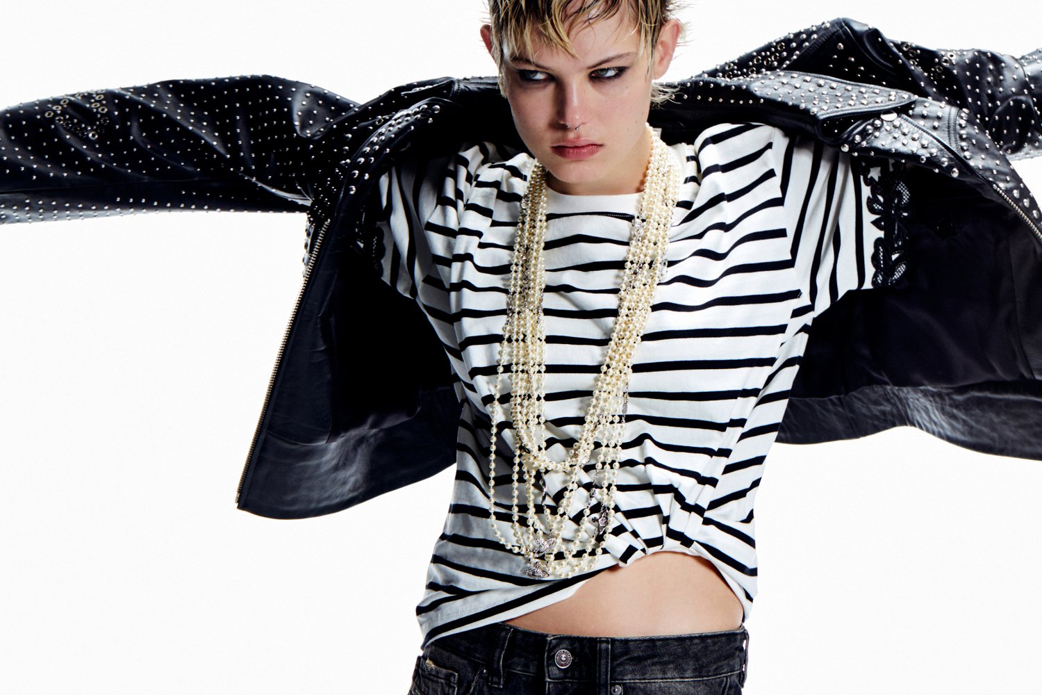 A model in a striped shirt and long pearl necklace pulls off a studded black leather jacket, showcasing an edgy fashion vibe.
