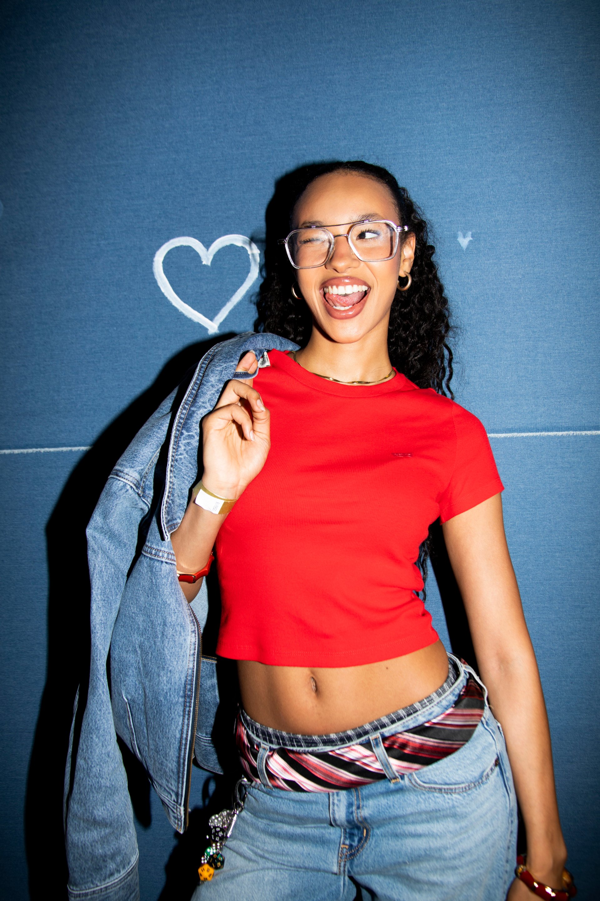 Rode t-shirt met korte mouwen, spijkerjasje over de schouder, blauwe jeans met een patroon in de tailleband. Achtergrond met een blauwe muur waarop een witte harttekening is.