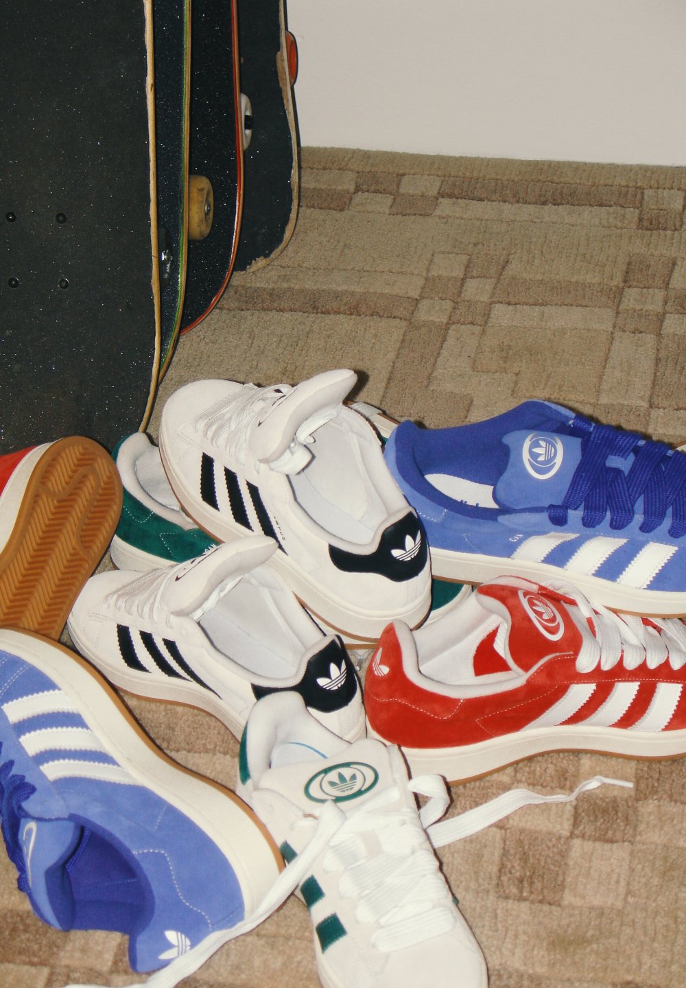 Farbenfrohe Auswahl an Adidas-Sneakers in verschiedenen Stilen, verstreut auf einem gemusterten Boden, neben einem Skateboard.