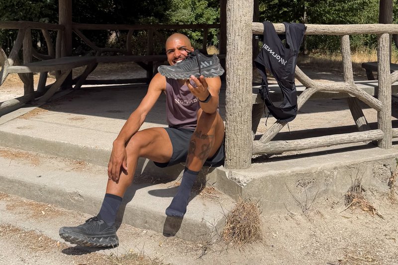 Man sitting on concrete step outdoors, holding up a black athletic shoe and smiling, with a wooden railing and backpack behind him.