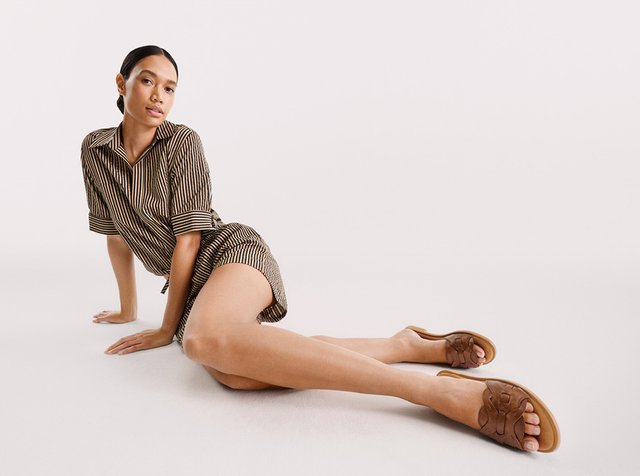 Frau mit dunklen Haaren, die ein gestreiftes, kurzärmeliges Kleid und braune Sandalen trägt und mit einem ausgestreckten Bein am Boden sitzt.