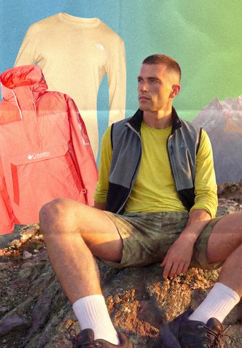 Muž v žltom tričku s dlhým rukávom a čiernej veste sediaci na skalnatom teréne, v pozadí červená bunda a béžová košeľa.