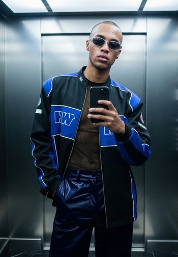 Giacca zip-up nera e blu con accenti bianchi, indossata sopra una maglietta in rete marrone. La modella indossa occhiali da sole sottili e ovali e pantaloni lucidi blu navy.
