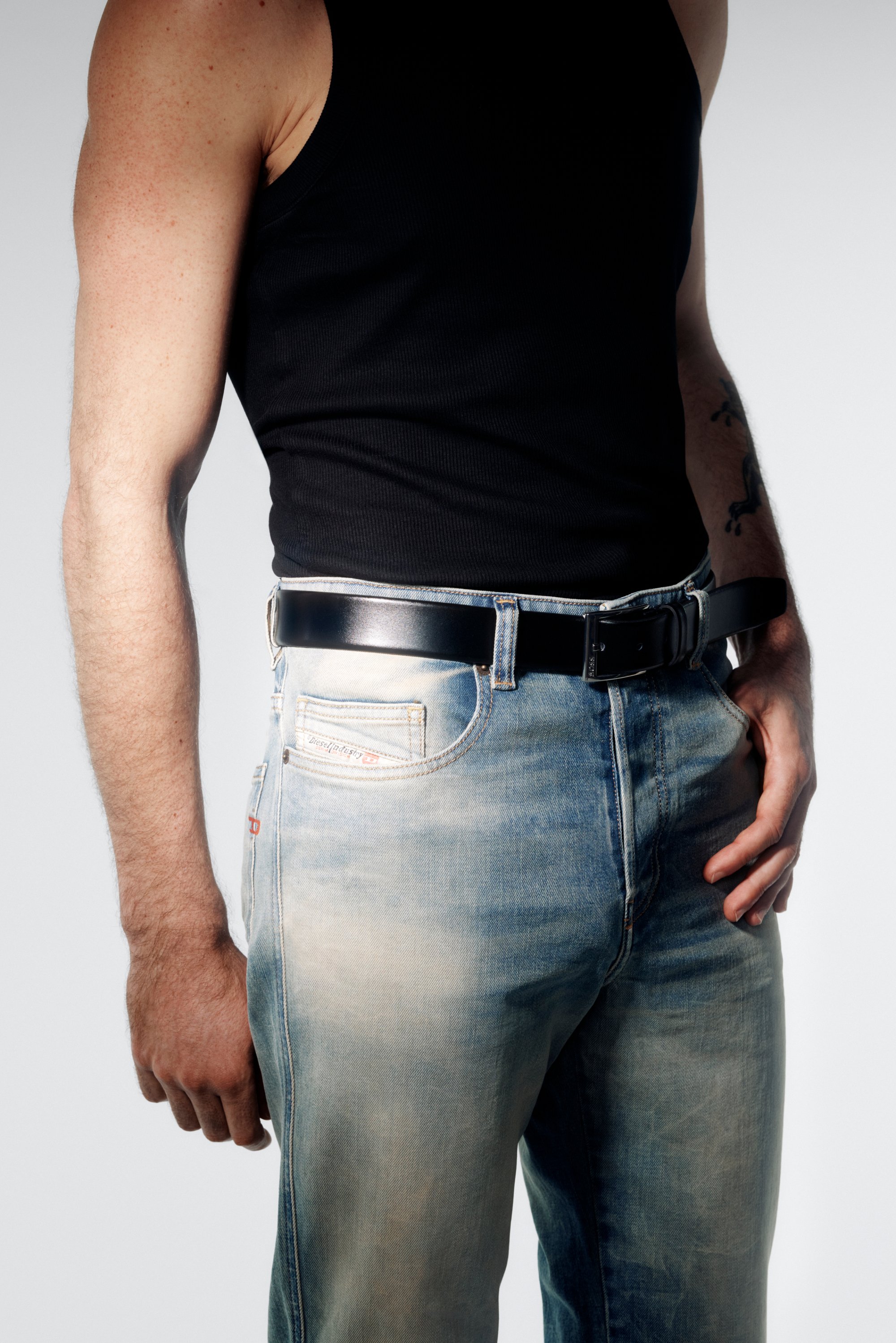 Una persona en una camiseta sin mangas negra está de lado, vistiendo unos jeans azules desgastados con un cinturón negro, mostrando un atuendo casual.