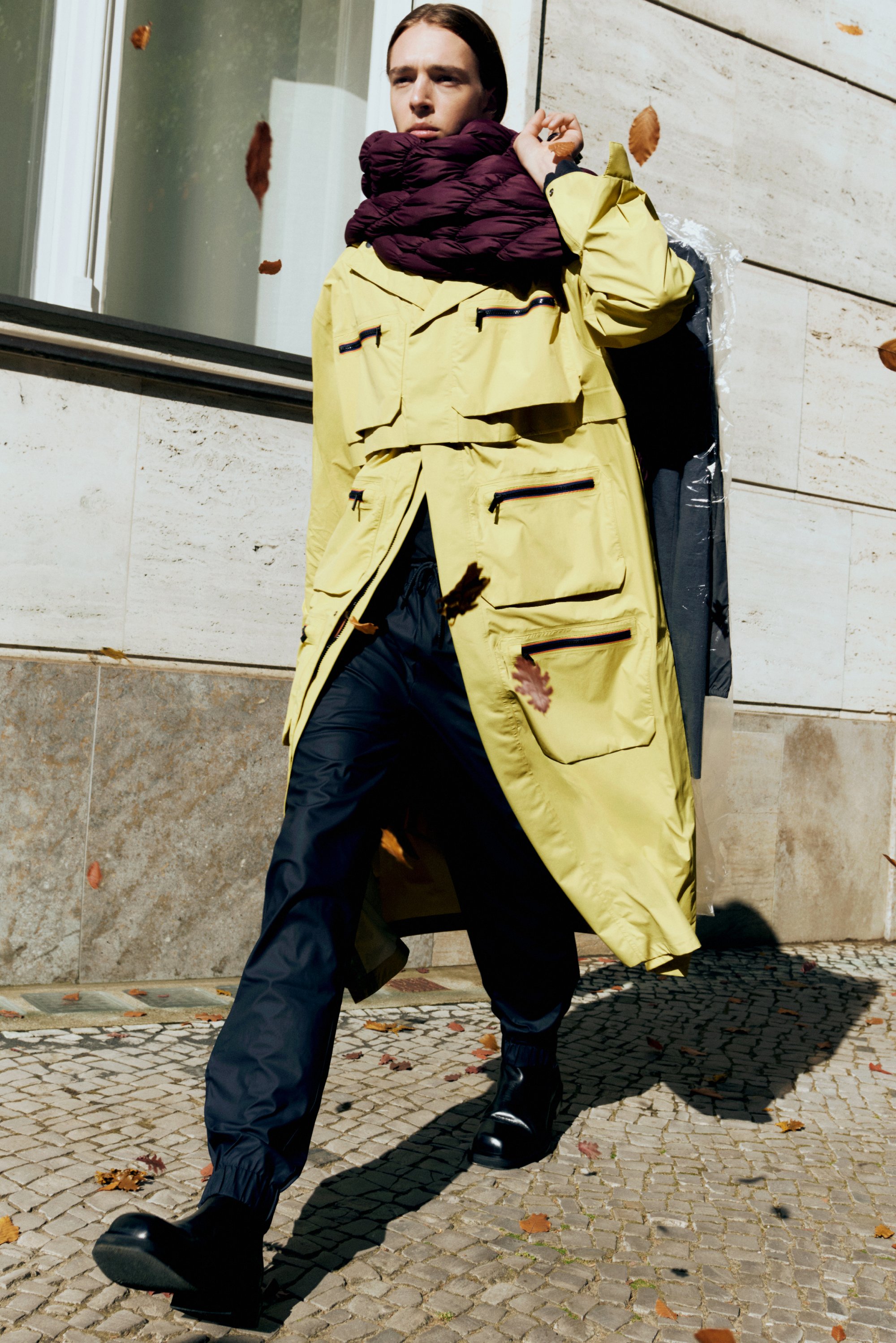 Gelber wasserdichter Mantel mit mehreren Taschen, kombiniert mit schwarzen elastischen Hosen und schwarzen Knöchelstiefeln. Ein lila Daunen-Schal rundet den Look ab.