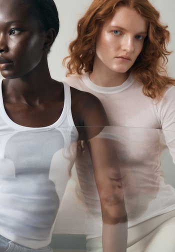 Twee modellen met verschillende huidtinten. Eén draagt een wit geribd tanktopje, de ander een licht doorschijnende lange mouw top. Zachte texturen, neutrale kleuren.