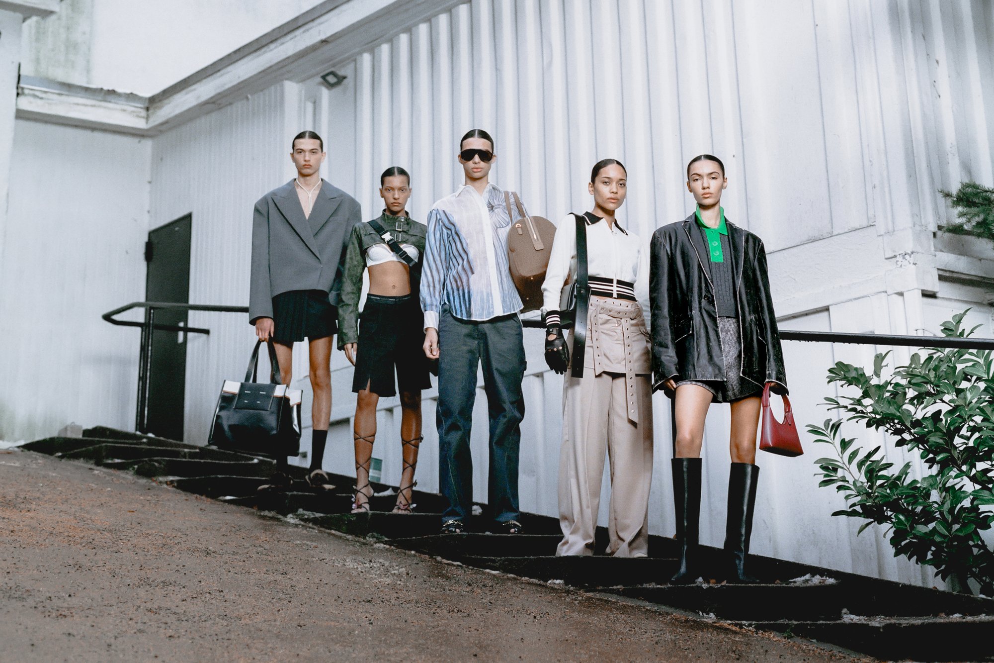 Five models pose on a staircase, showcasing various contemporary outfits and accessories, with a minimalist urban backdrop.