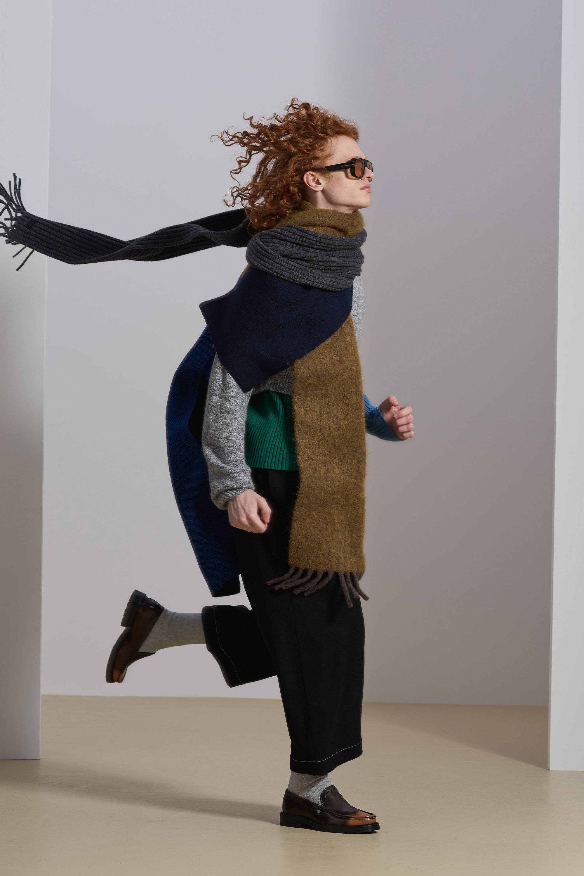 A person with curly red hair runs, wearing a layered scarf and oversized clothing, sunglasses, against a minimalistic background.