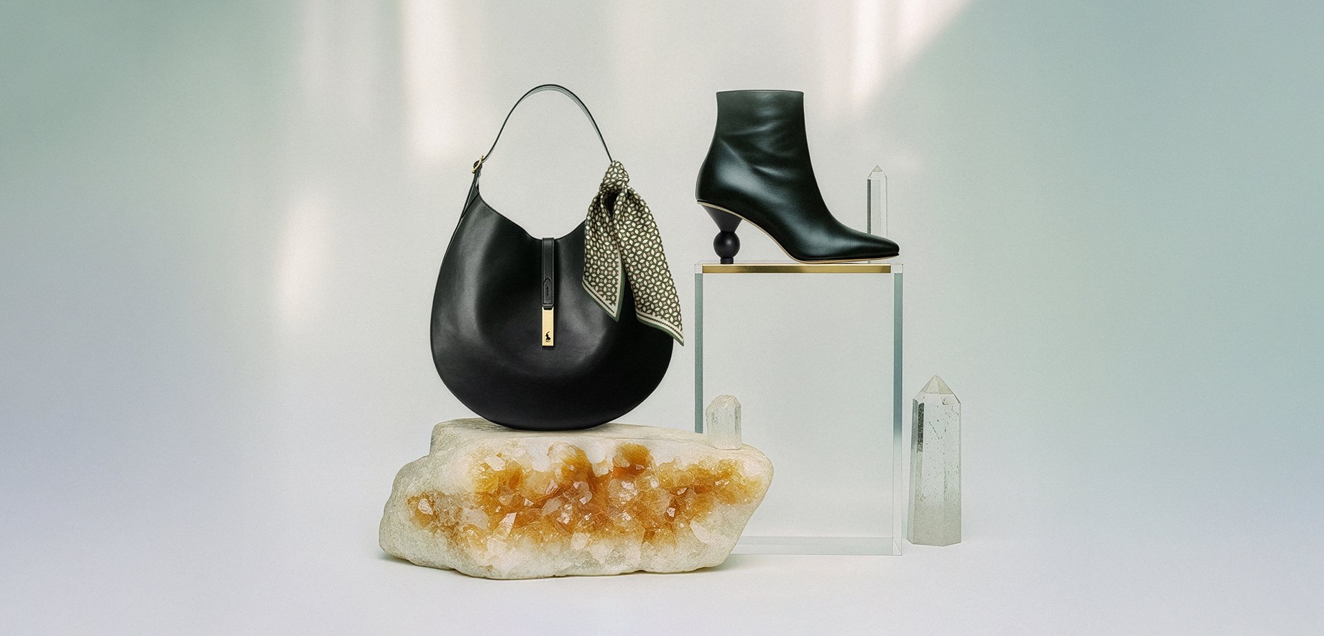 Black leather shoulder bag with a patterned scarf, matched with a black ankle boot featuring a distinctive sculptural heel, displayed on a crystal base.