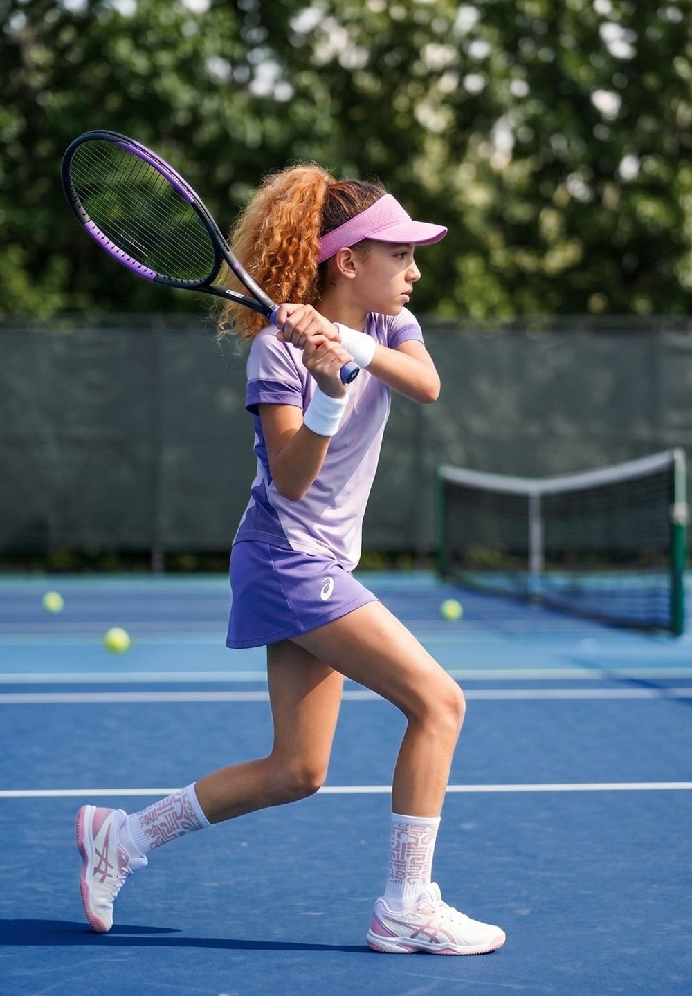 Young girl in purple sportswear poised to hit a tennis ball on an outdoor blue tennis court with tennis balls scattered around.